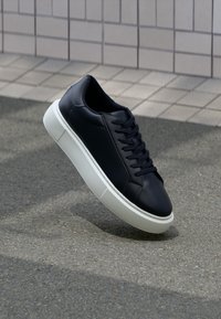 Schwarze Leder-Sneaker mit dicker weißer Sohle und schwarzen Schnürsenkeln, aufrecht auf einer gepflasterten Fläche neben einer gefliesten Wand positioniert.