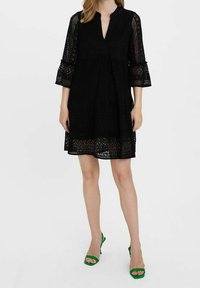 Femme portant une robe en dentelle noire longueur genou avec des manches cloche trois-quarts et des sandales à talons ouverts vertes.