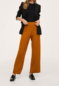 Pantalon à jambes larges jaune moutarde avec une texture côtelée et un ourlet effiloché, associ é à un haut noir ajusté et des mocassins foncés.