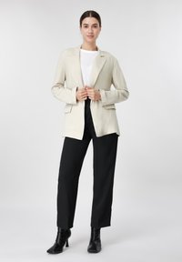 Femme portant un blazer beige, une chemise blanche, un pantalon noir et des bottines noires, debout devant un fond clair uni.
