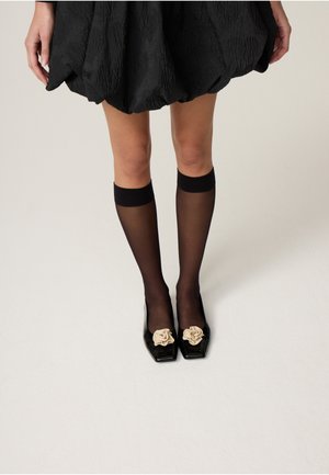 Legs wearing sheer black knee-high stockings and black square-toe shoes with beige flower decorations, paired with a black textured dress.