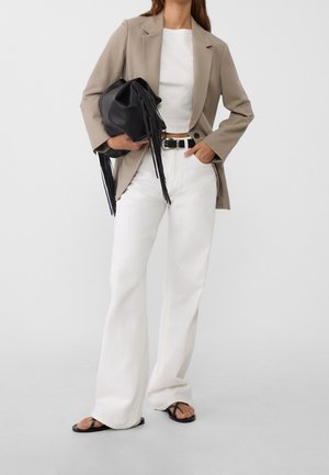 Beiger Blazer über einem weißen, bauchfreien Top, kombiniert mit weißen, ausgestellten Hosen und schwarzen Flip-Flops. Eine schwarze Tasche mit Fransenakzenten wird gehalten.