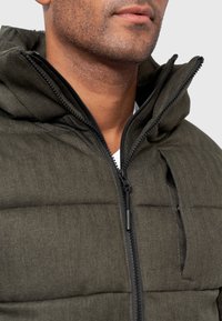 Parka s kapucí v tmavě zeleném herringbonovém vzoru; má plný černý zip a měkkou, texturovanou tkaninu.