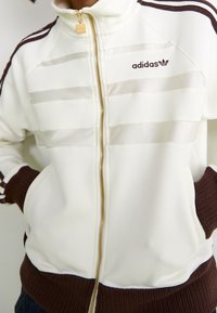 Adidas zip-jacka i krämfärgad med bruna accenter, med ribbade muddar och gulddragkedja, prydd med tre horisontella ränder.
