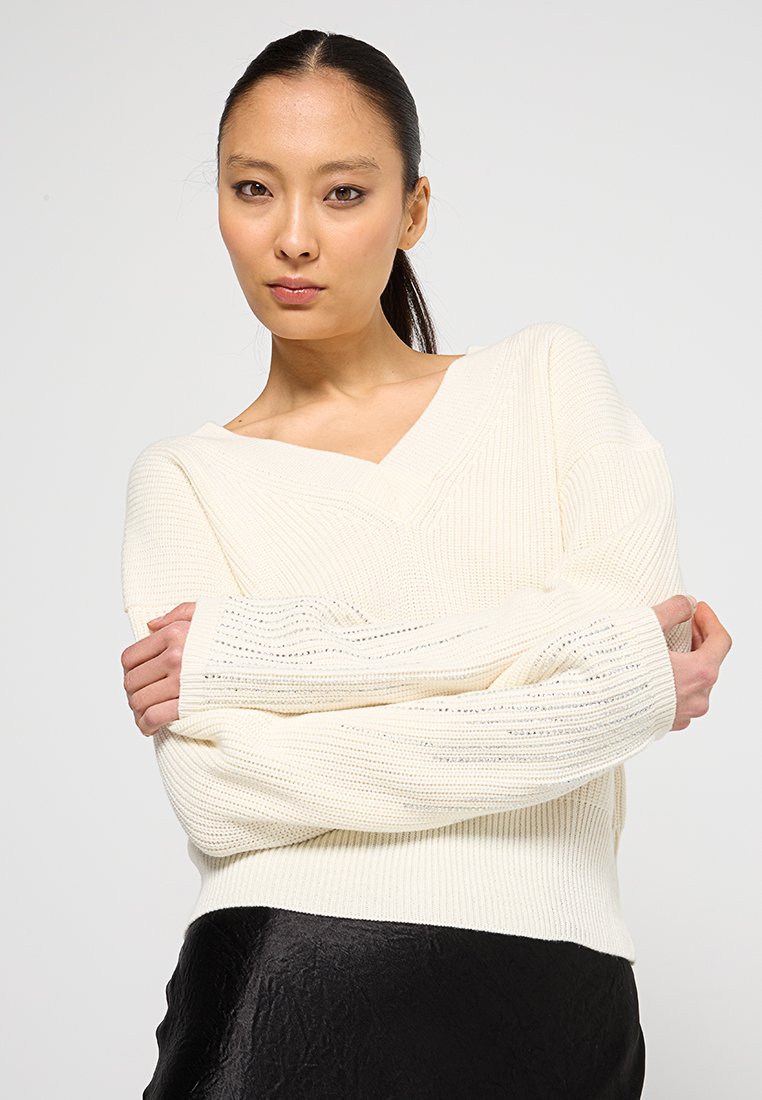 Maglione lavorato a maglia in crema con scollo a V, texture a coste e maniche lunghe. Indossato sopra una gonna nera lucida.