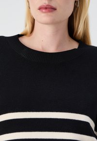 Črna sv sweater z rebrastim ovratnikom; ima bele horizontalne črte čez prsi, teksturirano pleteno tkanino in klasičen kroj.