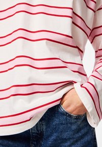 Person wearing a white long-sleeve shirt with pink and red horizontal stripes, hand tucked into blue denim jeans pocket.