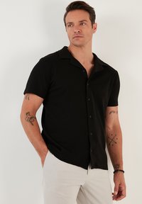 Camisa negra de manga corta con un tejido texturizado, cuello y botones al frente. Llevada por un hombre, combinada con pantalones de color claro.
