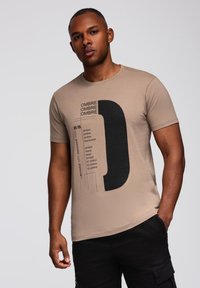 Beige puuvillane t-särk, millel on must graafiline tekst ja julge disainielement esiküljel. Ümmargune kaelus, lühikesed varrukad ja lõdva istuvusega.