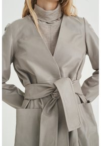 Jasnoszara skórzana płaszcz o długich rękawach, z paskiem w talii i dwiema przednimi kieszeniami. Gładka faktura i minimalistyczne szczegóły projektu.