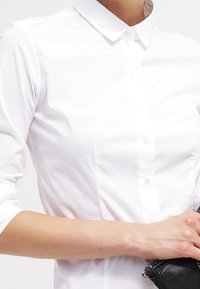 Camicia bianca con colletto, maniche lunghe e design aderente, realizzata in morbido cotone e con pieghe sottili.