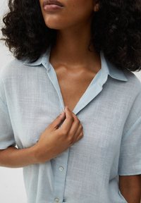 Camicia azzurro chiaro a maniche corte con una leggera lucentezza, colletto abbottonato, tessuto testurizzato e dettagli di cucitura visibili. Mano che aggiusta il colletto.