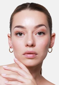 Boucles d'oreilles en or avec une finition texturée, présentant un design circulaire. La peau lisse et le maquillage léger mettent en valeur les traits du visage.