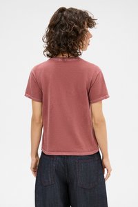Short-sleeve, relaxed-fit T-shirt in muted red cotton fabric. Features a crew neckline and slightly cropped hem, worn with dark jeans.