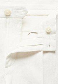 Gros plan sur la ceinture d’un pantalon blanc déboutonnée, avec le bouton et une bande élastique réglable intérieure visibles.
