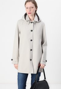 Trench coat impermeabile grigio chiaro con cappuccio, dotato di bottoni neri e dettagli ai polsini. Indossato sopra jeans blu, abbinato a una borsa nera.