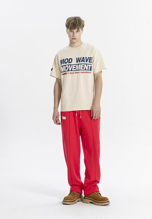 Joven de pie con una camiseta beige "MOD WAVE MOVEMENT", pantalones rojos holgados y zapatos marrones sobre un fondo blanco.