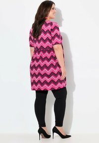 Růžovo-černá tunika se vzorem cik-cak, krátké rukávy, v kombinaci s černými legínami a černými vysokými podpatky. Hladký materiál a volný střih.