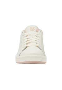 Zapatilla de deporte baja en blanco y rosa claro con cordones, laterales perforados y un logo de escudo en la lengüeta, vista desde el frente.