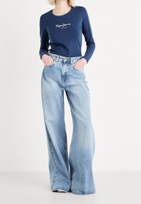 Marinblå långärmad skjorta med vitt logotyp, kombinerat med vida ljusblå jeans. Båda plaggen har klassiska stilar utan ytterligare mönster.