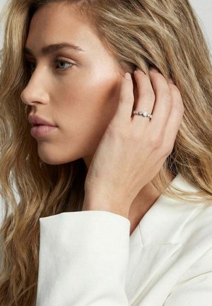 Ung kvinde med løsthængende blondt hår iført en hvid top, der holder hånden tæt ved øret og viser en ring med runde ædelsten.