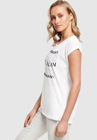 White cotton T-shirt with rolled short sleeves featuring black text reading "BERLIN GERMANY" in varying fonts and sizes. Smooth texture.