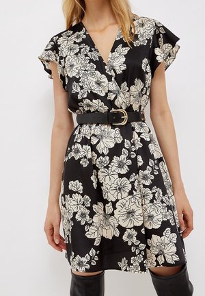 Femme portant une robe portefeuille noire avec un grand imprimé floral blanc, des manches courtes, et une large ceinture noire avec une boucle dorée.