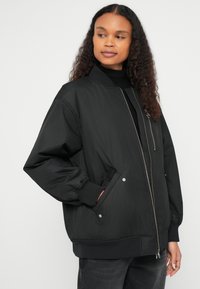 Μαύρο μπουφάν τύπου bomber με κλείσιμο με φερμουάρ, ριμπ μανσέτες και δύο πλαϊνές τσέπες. Κατασκευασμένο από απαλό, ελαφρύ υλικό με άνετη εφαρμογή.