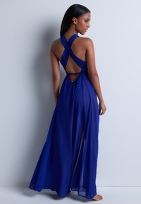 Robe maxi en chiffon bleu avec un dos entrecroisé, présentant une découpe et une silhouette fluide, offrant un aspect aérien et élégant.