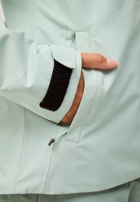Giacca verde menta con un materiale esterno liscio, polsini regolabili in Velcro nero, mano in tasca laterale e dettaglio del logo sulla manica.