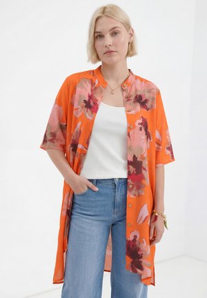Blonde woman wearing an orange floral buttoned shirt over a white tank top and blue jeans, standing with one hand in pocket.