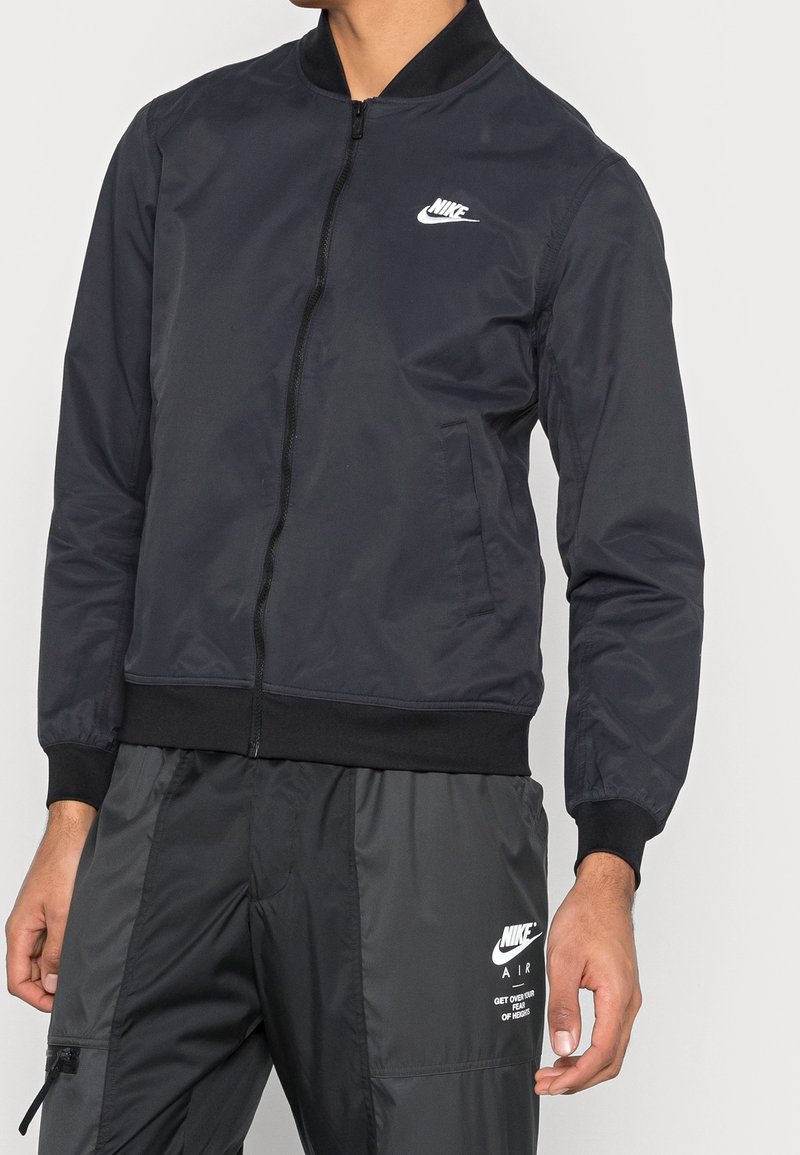 Zwarte nylon bomberjack met ritssluiting, geribbelde manchetten en zoom, met een wit Nike-logo op de bovenste linkerborst.