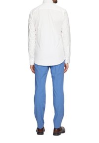 Witte overhemd met lange mouwen en een klassieke kraag, gecombineerd met lichtblauwe op maat gemaakte broek en bruine schoenen, gezien van achteren.