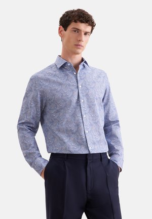 Mann mit lockigem Haar trägt hellblau gemustertes langärmeliges Hemd und dunkle Hose, steht mit den Händen in den Taschen vor weißem Hintergrund.