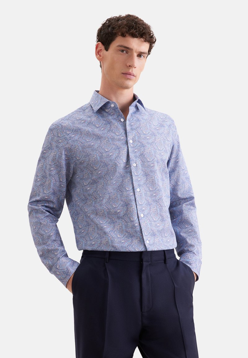 Homme aux cheveux bouclés portant une chemise à manches longues bleu clair à motifs et un pantalon foncé, debout les mains dans les poches devant un fond blanc.