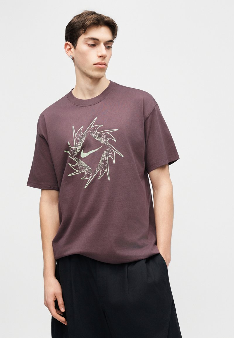 Junger Mann trägt ein violettes Nike T-Shirt mit einem stilisierten Swoosh-Logo und schwarzen Hosen, steht vor einem schlichten hellen Hintergrund.