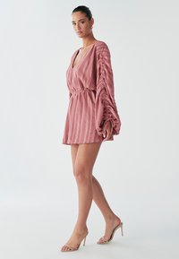 Model trägt ein pinkes, plissiertes Kleid mit wehenden Ärmeln, kombiniert mit Riemchen-High-Heels, und steht selbstbewusst vor einem hellen Hintergrund.