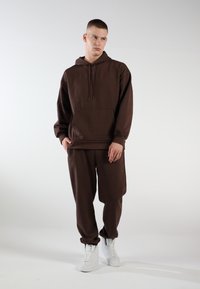 Brauner Kapuzenpullover mit einer Vorderseite Tasche und gerippten Bündchen, kombiniert mit passenden Jogginghosen.getragen mit weißen Turnschuhen. Einfacher, weicher Stoff.