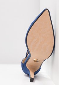 Escarpin bleu en daim avec un bout pointu, doté d'une semelle en cuir beige et d'un accent logo doré près du talon.