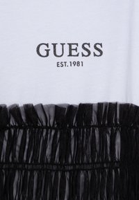 Țesătură albă ce prezintă "GUESS EST.1981" brodat în negru, cu o margine din tul negru plisat care adaugă textură și contrast vizual.