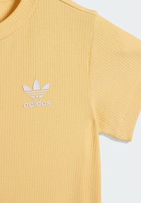 Żółta prążkowana koszulka wykonana z miękkiej tkaniny, z białym logo Adidas na piersi oraz trzema akcentami w kształcie liści nad nazwą marki.