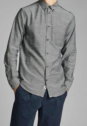 Mann trägt ein graues Hemd mit Brusttasche und dunkelblaue Hose, Hände in den Taschen, steht vor einem einfarbigen Hintergrund.