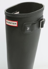 Juoda guma lietaus batai su apvaliu viršumi, su sagties detalėmis ir raudona bei balta prekės ženklo etikete, pažymėta "HUNTER".