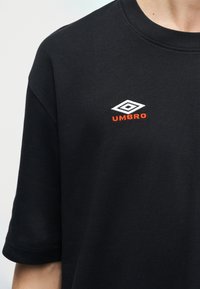 Μαύρο κοντομάνικο πουκάμισο που φοριέται από άτομο, με λευκό διπλό διαμαντένιο λογότυπο και κείμενο "UMBRO" κεντημένο σε πορτοκαλί χρώμα στο στήθος.