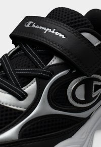 Chaussure de sport noire avec une tige en mesh, des accents argentés et une bande velcro proéminente. Comprend des lacets épais et une semelle rembourrée.