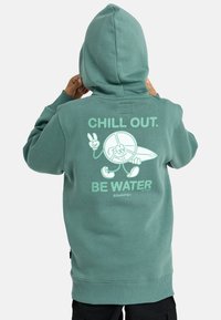 En person med en teal hoodie med huva upp, som visar baksidan med text och grafik av en fredssymbolkaraktär som håller en surfbräda med texten "CHILLA. VAR VATTEN."