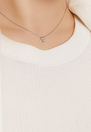 Collier en chaîne d'argent avec un petit pendentif triangulaire porté sur un pull blanc à col en V côtelé.