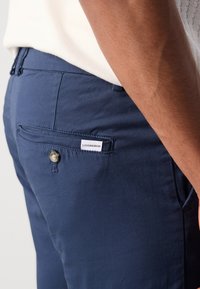 Pantalon bleu marine en tissu lisse, doté d'une poche arrière avec fermeture à bouton et d'une étiquette blanche portant le nom de la marque "LINDBERGH."