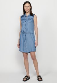 Abito di jeans con scollatura a colletto, design senza maniche, bottoni frontali, due tasche sul petto, vita regolabile con coulisse e dettagli blu.
