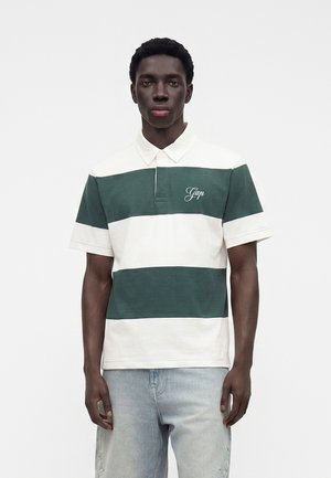 Jeune homme portant un polo rayé vert et blanc avec le logo "Gap" et un jean bleu clair, debout devant un fond blanc uni.